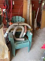 Four stacked green resin Adirondack chairs in a covered storage area. Two chairs are clearly visible with a few garden hoses on top of them. Chairs have surface dirt and scratches.