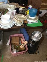 Photo showing a collection of white and patterned china and Corelle dishes, ceramic mugs, plastic food covers stacked on and around a small table, with a pink plastic tote holding various toiletry products and boxes below, as well as the Bionaire air cleaner nearby on the floor.
