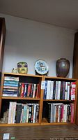 Pair of wood bookshelves with assorted books, porcelain urn, decorative plate, glitter mask, and stone lighter on top shown against basement wall