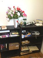Two shelves showing various items including books, cameras, massager, faux flowers, and battery lantern.