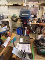 Front view of the blue and black MasterCraft branded drill press alongside the drill bits box and instruction manual on a wood surface.