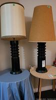 Two tall ceramic lamps with black ribbed bases; one has a white fabric shade with lace trim, the other has a plain brown jute shade. Lamps placed on separate small tables against a neutral wall.