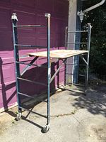 Front side view of single level rolling scaffold with wooden platform and caster wheels, showing structure and wear.