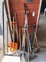 Full view of various gardening tools leaning against a red wall including shovels, rakes, demolition bars, and handles visible.
