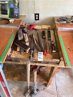 Photo showing assortment of wire brushes, metal files, and a small hand planer, laid out on a wooden workbench with sledgehammers underneath.