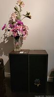 Pair of black fabric-covered speakers with artificial flower arrangement in clear glass vase placed on top, showing overall lot setup.