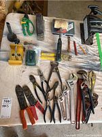 Overhead view of assorted hand tools spread on white cloth including cutters, wrenches, pliers, pruner, and digital device
