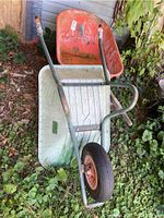 Two wheel barrows stacked on each other outdoors in a grassy and leafy area. One barrow has a light green metal basin and the other an orange resin basin.