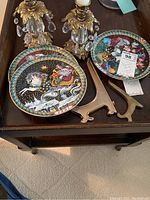 Photo showing two Christmas decorative plates with Santa Claus and reindeer sleigh designs, two metal plate holders, and one gold tone candleholder.
