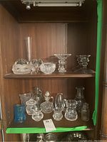 Two shelves of cut glass including candleholders, miniature pitchers, miniature vases in clear faceted and frosted designs.