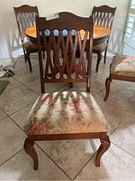 Front view of one wooden dining chair with floral upholstered seat and carved lattice backrest, four chairs visible in background around a wooden dining table.