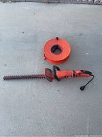 Full view of the orange hedge trimmer with cord and manual cord reel on concrete surface.