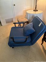 Blue folding chair cot with black armrests and blue pillow shown in chair form near a side table and chair.