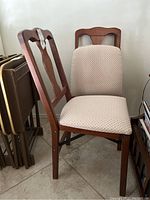 Three wooden folding chairs with padded cloth seats stacked next to folded tables, showing front and side view.