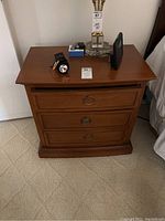 Front view of one wooden night stand with three drawers and round metal ring pulls, medium-brown finish, placed on carpet next to baseboard and wall