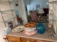 Countertop with stacked platters including porcelain and wooden platters, blue glass vase with peacock feather, and seven wine glasses on shelves.