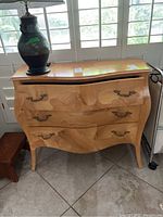 Front view of a wood side table with curved front and three drawers with brass handles. Light colored wood with visible grain patterns.