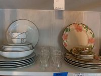 Stack of white Noritake China dinnerware with silver trim, including plates and cups, visible glassware behind.