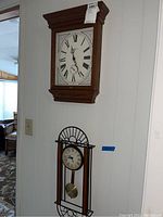 Two Howard Miller wall clocks displayed on a white wall; one large wooden rectangular clock and one smaller clock with glass front and pendulum.