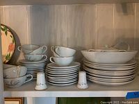 Photo showing stacks of white china cups, saucers, plates, salt and pepper shakers, and large serving bowl with gold floral pattern