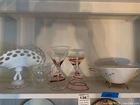 Shelf with milk glass compote, clear glass cups, martini glasses with red swirl pattern, and a white ceramic bowl with gold stand and lid.