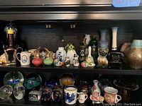 Wide shelf view of mixed ceramic figurines and glass paperweights, arranged in rows on dark wood shelving.