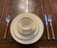 Full place setting with stacked plates, cup and saucer, flanked by spoon, knife, and fork with brown handles on wood table