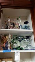 View of all glassware items on two shelves inside cabinet, showing pitcher, plates, and various stemware pieces with colors blue, green, and clear glass.