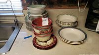 Stack of bowls and plates including white bowls with aqua tint and ribbed texture, plain white plates with colored rims, decorative maroon plate with floral patterns, and beige plates with brown line detail.