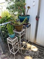Front view showing all three metal plant stands and three potted plants on top, arranged by size, outside next to a house and chain fence.