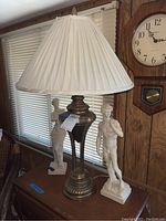Photo showing bronze table lamp with pleated white shade and two white classical statue figurines standing on a table in front of window blinds.