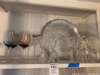 Full set showcasing 10 assorted colored glass stemware glasses alongside a large round decorative metal platter with scalloped edges, displayed on a white shelf.