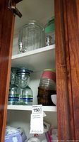 Wide angle view of cabinet shelf showing large glass beverage dispensers, stacked cups with colored rims, and stacked bowls of various colors and sizes.