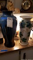 Two vases placed on a countertop, one tall dark blue vase with vertical ridges and one white ceramic vase with green ivy leaf designs and purple shading.