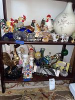Wide view of glass and ceramic figurines arranged on two shelves including roosters, ducks, small decorative pieces, and large white ceramic vase in background.