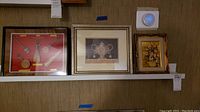 Overview of three framed wall art pieces hung on a wall including musical instrument illustration, urn artwork and floral painting.