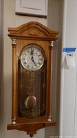 Full view of the wooden pendulum wall clock with glass front and decorative top and pillars.