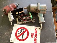 Two air impact wrenches, assorted machinist tools, and a No Smoking sign on a black surface.