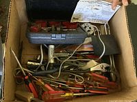 Box filled with assorted vintage hand tools including screwdrivers, pliers, and hole saw set.