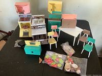 Wide shot showing entire set of pastel-colored doll kitchen furniture, chairs, table, and bags of plastic food accessories on a black surface.