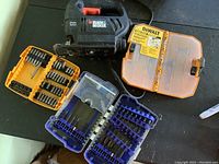 Photo showing a Black & Decker jigsaw, two cases with assorted drill bits, and an orange DEWALT plastic tool storage box on a black surface.