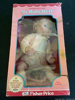Front view of the Fisher Price My Baby Beth Doll in original pink box with clear window showing the doll wearing a cream knit cardigan, white undershirt, floral diaper cover, and matching booties.