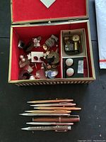 Top view of an open box with multiple costume jewelry pieces including brooches, pins, and earrings, with a set of Cross fountain pens laid out in front of the box.