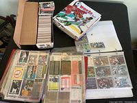 Photo showing a cardboard box filled with loose sports cards, a stack of loose cards on the table, a sports card album, and three plastic pages filled with cards.