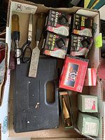Overview of a cardboard box containing fishing line spools, hand tools with wooden handles, vintage light bulb boxes, and a closed black plastic carrying case.