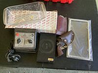 Photo shows six assorted items on black surface including Hornby transformer, glass tray, leather toy gun holster with toy revolver, wallet, and metal-framed glass plate.