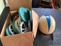 Box with many vintage movie film reels in metal canisters and a vintage motorcycle helmet beside it