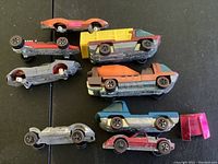 Photo of seven vintage Hot Wheels cars on black surface showing redline wheels and various colors and models.