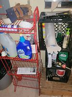 Photo showing red metal wire shelf with gloves and cleaning supplies beside three stacked black milk crates holding various bathroom and cleaning products.