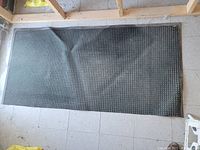 Full view of the black heavy duty floor mat laid flat on a tiled floor showing its rectangular shape, textured pattern, and wrinkling.
